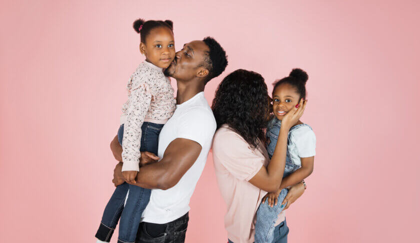 A happy family dressed in Jeans and white and pink t-shirts smiles and kisses one another.