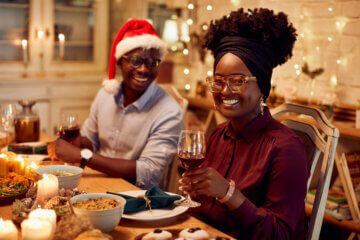 A woman sits at a table during a holiday party. The table is lit with candles and twinkle lights. She holds a glass of wine alongside her friend, who is wearing a Santa hat. They are filled with holiday cheer.