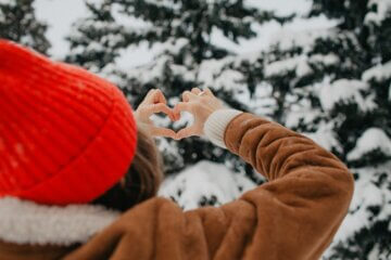 A woman is seen outdoors making a heart with her hands. Beyond her hands is a pine tree, coated with snow. The woman is wearing a tan coat and red winter hat.