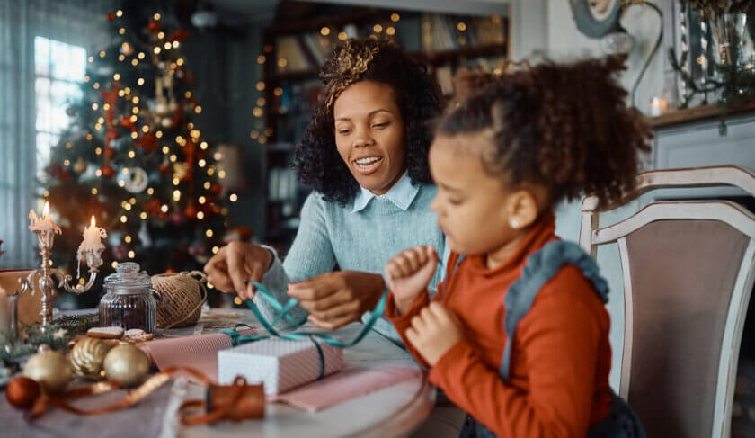 A mother and young daughter are seated at a table wrapping gifts. There is a Christmas tree in the backgroung.