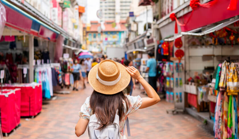 A woman is seen walking through a street market. She is wearing a wide brimmed hat.