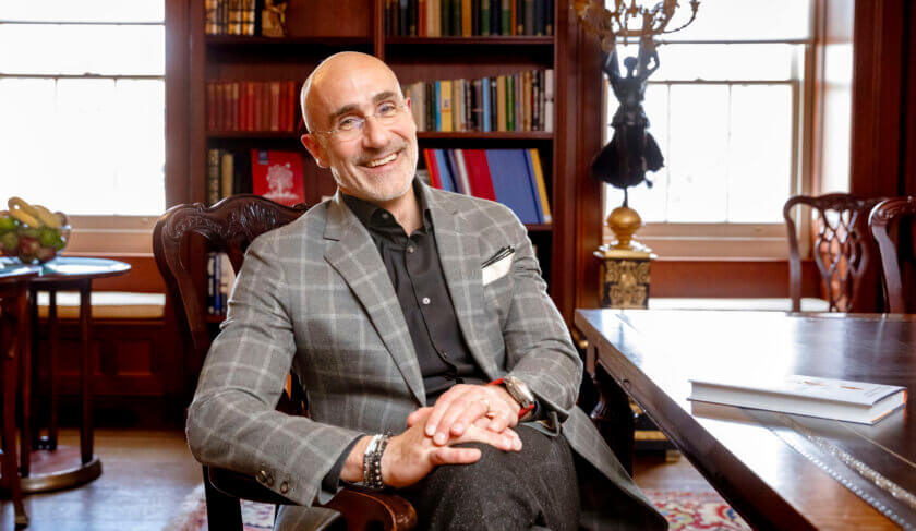 Arthur Brooks wears a suit and smiles at the camera, relaxed in a chair with books in the background.