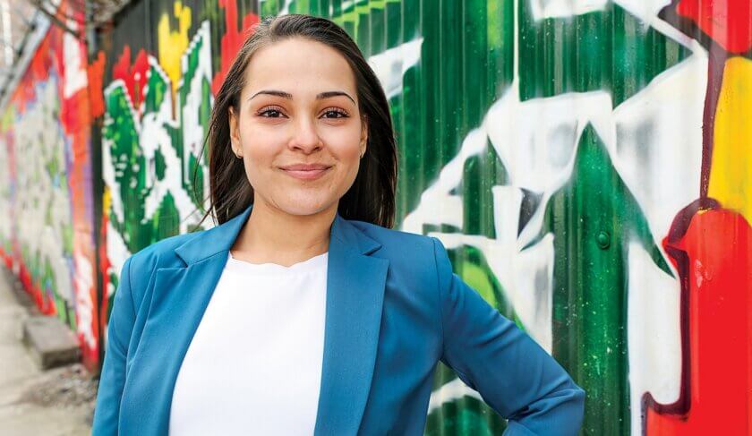 Headshot of Yanely Espinal wearing a blue blazer with graffiti behind her outside.