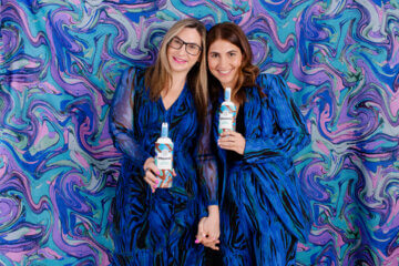 The sisters who founded the whipped cream company Whipnotic pose against a blue and purple background with their product