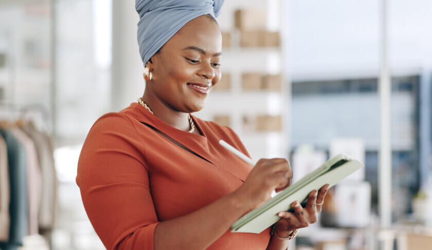 A smiling woman wearing a blue headscarf, gold earrings and a red clay-colored dress writes down her New Year goals in a notebook and contemplates her success.