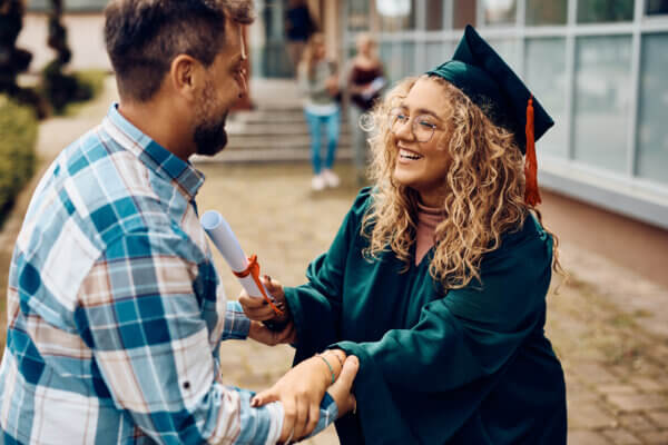 A college graduate with blonde curly hair and glasses embraces her father. He is wearing a plaid shirt and has a beard. They both look very happy.