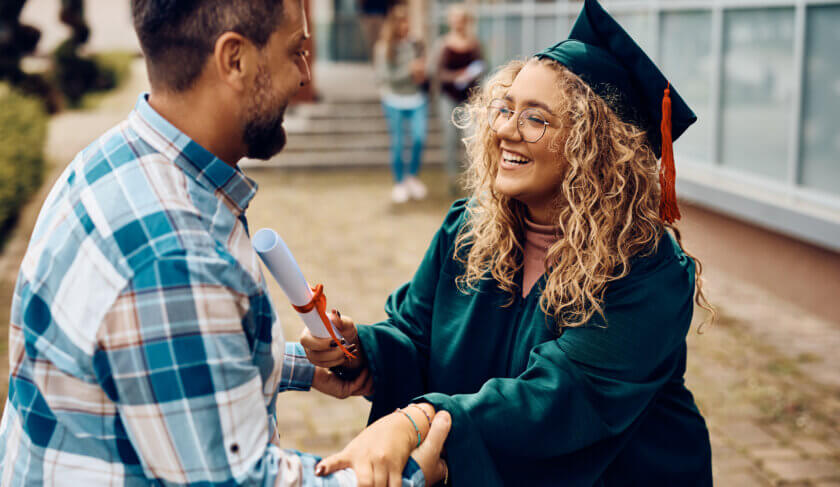 A college graduate with blonde curly hair and glasses embraces her father. He is wearing a plaid shirt and has a beard. They both look very happy.