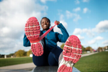 A woman laces up pink running shoes and smiles against a bright blue sky and green grass. She wears a blue long-sleeve pullover and black pants and is ready to go for a run on a neighborhood sidewalk.
