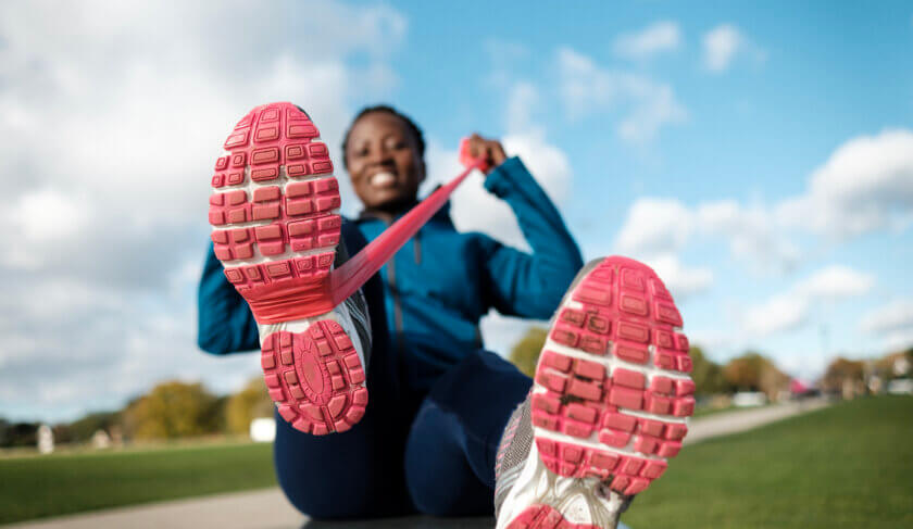 A woman laces up pink running shoes and smiles against a bright blue sky and green grass. She wears a blue long-sleeve pullover and black pants and is ready to go for a run on a neighborhood sidewalk.