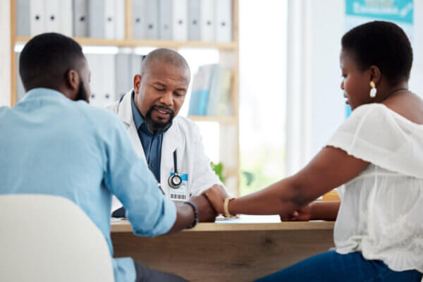 A young black couple is seen sitting at a desk and holding hands. They are talking to a doctor.