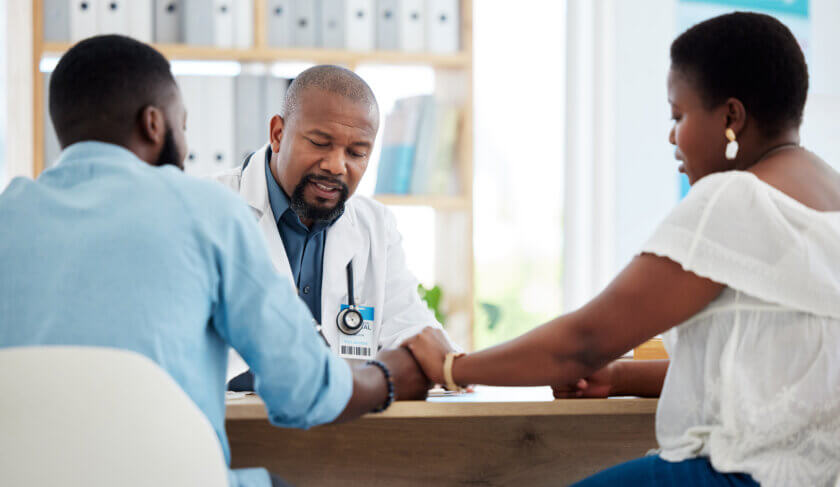 A young black couple is seen sitting at a desk and holding hands. They are talking to a doctor.