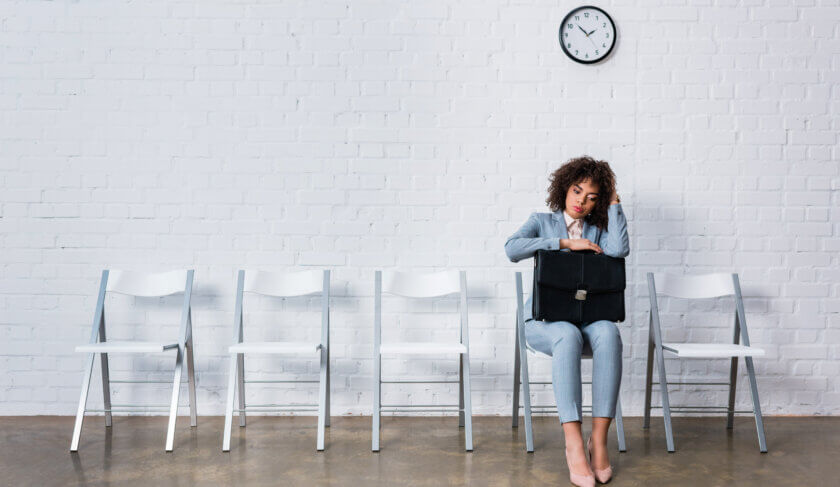 A woman in a blue business suit holds a briefcase and sits along a white wall with five empty chairs. She waits for a job interview.