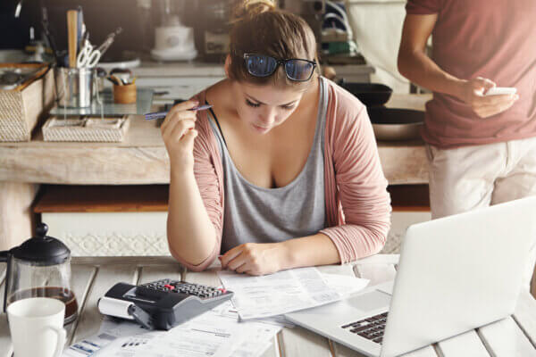 A woman sits at her kitchen counter with a laptop, paperwork and a calculator and does taxes. She wears a pink top and has glasses on top of her head, and has a cup of coffee next to her.