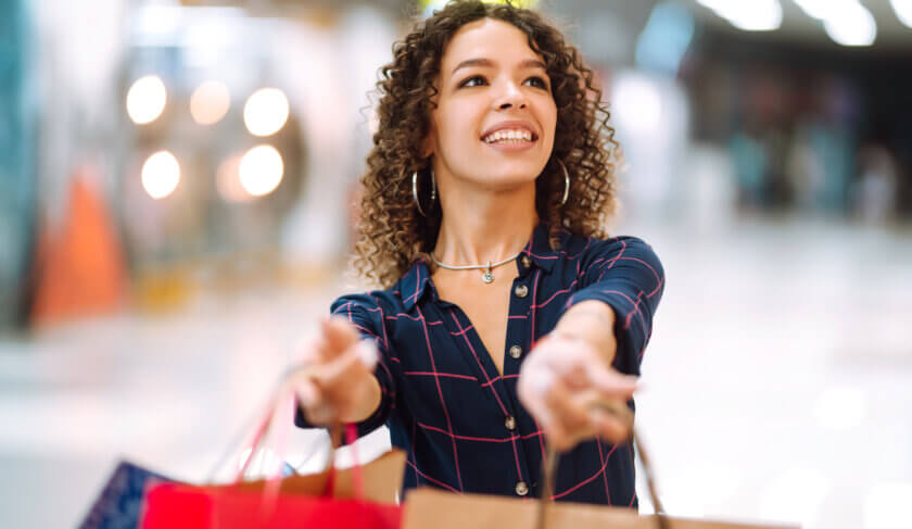 A beautiful woman with curly hair and a navy checkerboard shirt on smiles as she holds out shopping bags. She has been budgeting and learning how to spend smartly.