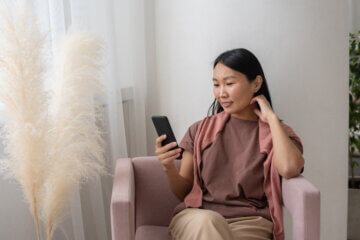 A brunette woman wearing a pink top sits in a pink chair and checks her budgeting app on her phone as she continues her journey to become a frugal money saver. She sits in an all-white room with white flowers in a vase and relaxes in the chair.