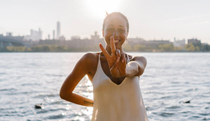 A beautiful woman wearing a yellow tank top stands on the water's edge along the East River in Brooklyn. She smiles and flashes a peace sign.