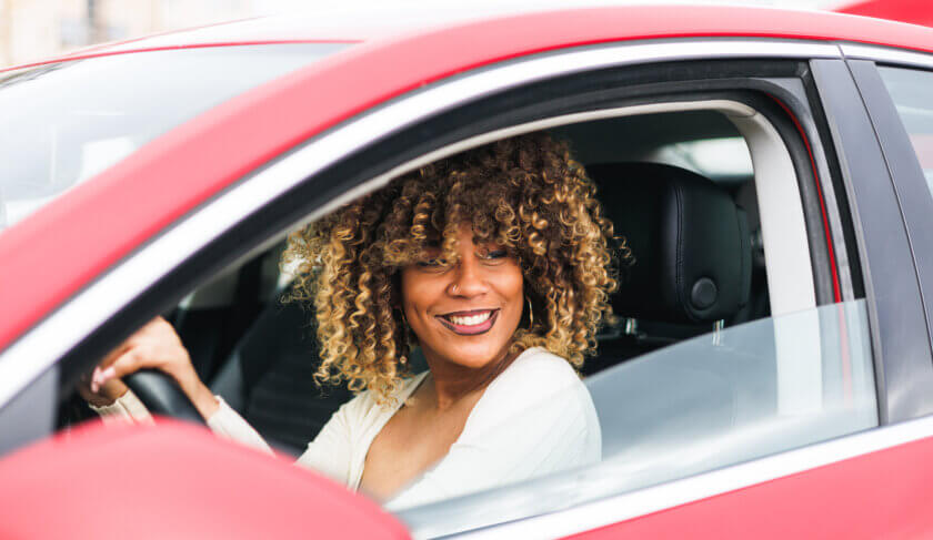 A woman is sitting behind the wheel of a red car. She is black, with curly hair and wearing a white top. She is smiling.