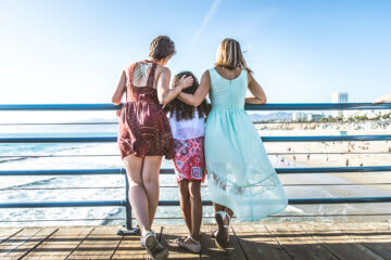 A mother stands along a beach boardwalk with her daughters. They wear colorful dresses and look out at the water