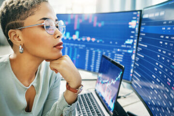 A professional woman wearing a seafoam green blouse looks at a double monitor displaying a stock market ticker, as she evaluates financial risk in the market.