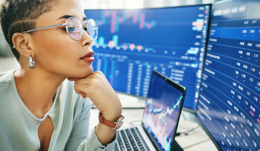 A professional woman wearing a seafoam green blouse looks at a double monitor displaying a stock market ticker, as she evaluates financial risk in the market.