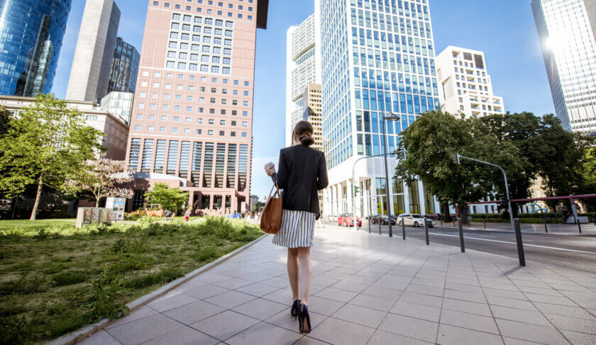 A professional woman walks through downtown Frankfurt amongst skyscrapers and blue sky.
