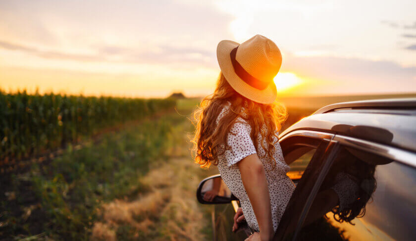 A happy woman on a summer road trip leans out her car window towards the sunset.