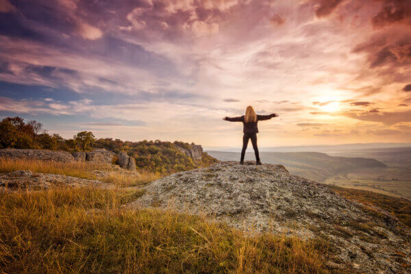 A woman stands at the top of a mountain along the ocean, against a beautiful multi-colored sunset