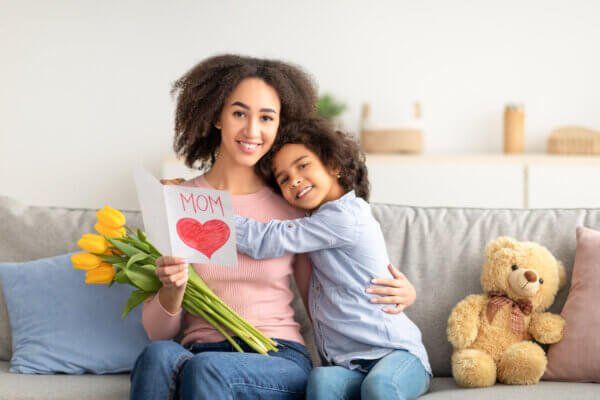 A mother and daughter are sitting on a couch. The daughter has given her mom a bouquet of tulips and a card.