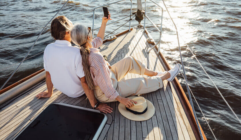 Two seniors sitting together on a yacht bow and taking selfie at sunset as their boat floats in the ocean.