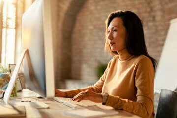 An Asian woman is seated at a desk looking at a computer.