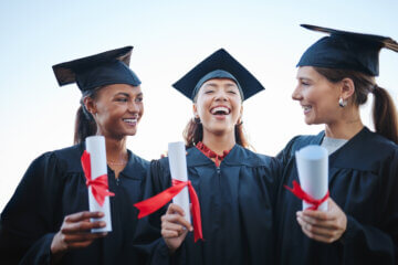 Three friends are seen post-college graduation talking and laughing with one another. They are wearing their caps and gowns and holding diplomas.