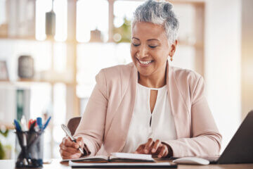 A woman with gray hair is seen sitting at a desk writing in a notebook. She appears happy.
